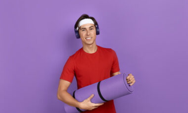 Junger lächelnder Sportler oder Trainer hält eine Fitness- oder Yogamatte in den Händen, er trägt ein rotes T-Shirt, blaue Kopfhörer und ein weißes Stirnband, isoliert auf violettem Hintergrund.