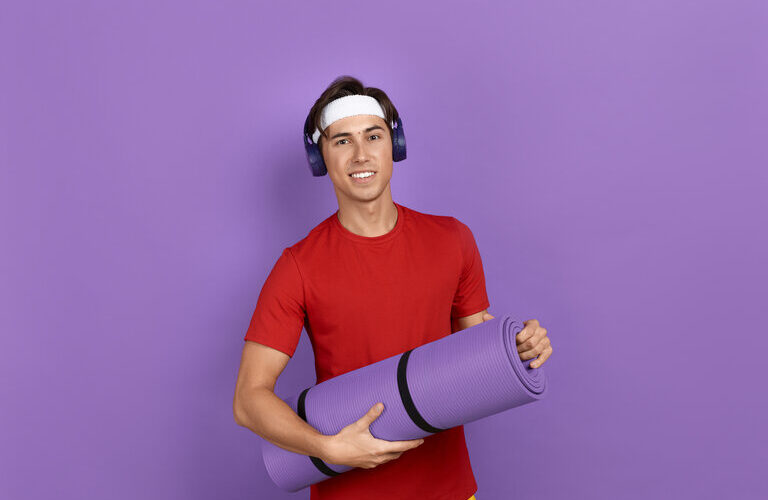 Junger lächelnder Sportler oder Trainer hält eine Fitness- oder Yogamatte in den Händen, er trägt ein rotes T-Shirt, blaue Kopfhörer und ein weißes Stirnband, isoliert auf violettem Hintergrund.