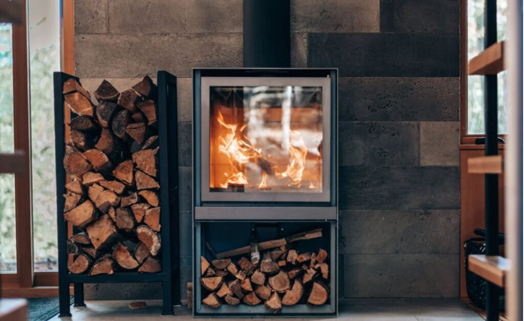 Ein moderner Kamin mit klarer Glasscheibe für die Kaminofentür, umgeben von Holzvorräten, in einem stilvoll eingerichteten Raum. Perfekt für gemütliche Abende.