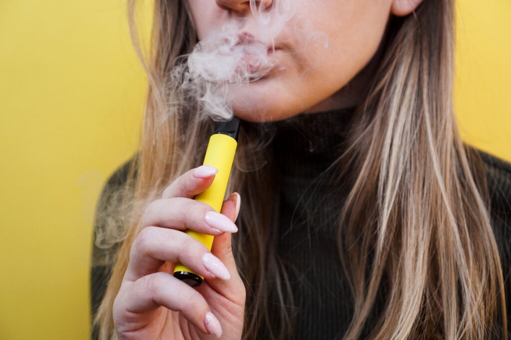 Eine junge Frau verwendet ein wiederverwendbares Vaping-Gerät wie Elfbar auffüllbar. Sie hält das Gerät vor ihrem Gesicht und inhaliert Dampf, was die moderne und nachhaltige Alternative zu herkömmlichen Rauchmethoden symbolisiert.