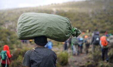 Träger trägt große Last auf dem Kopf während der Besteigung des Kilimandscharo, mit einer Wandergruppe im Hintergrund.