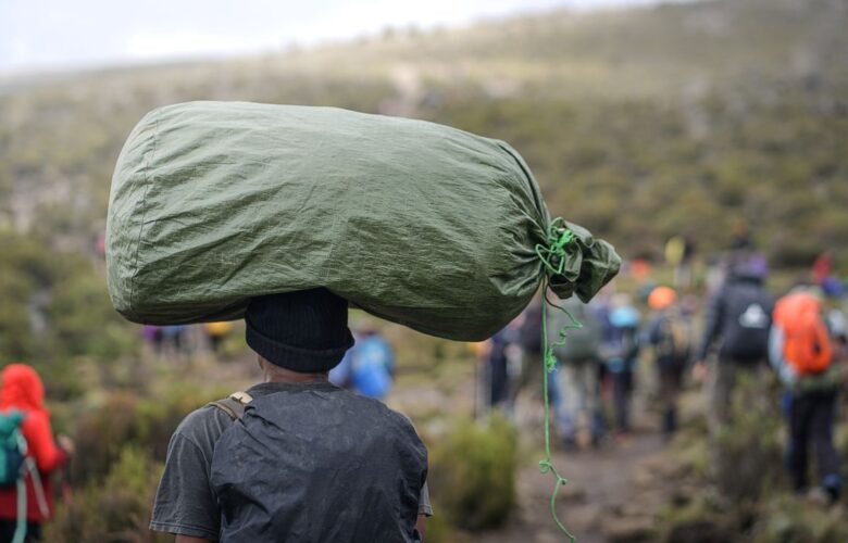 Träger trägt große Last auf dem Kopf während der Besteigung des Kilimandscharo, mit einer Wandergruppe im Hintergrund.