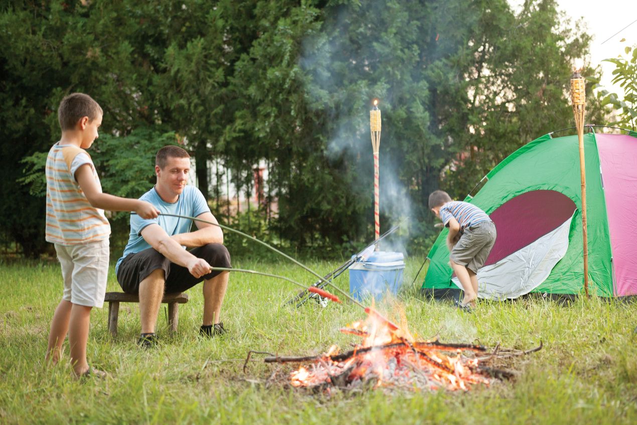 Vater grillt mit seinen Kindern Stockbrot über einem Lagerfeuer, während ein grünes Zelt im Hintergrund steht – die Campingtoilette sorgt für Hygiene unterwegs.