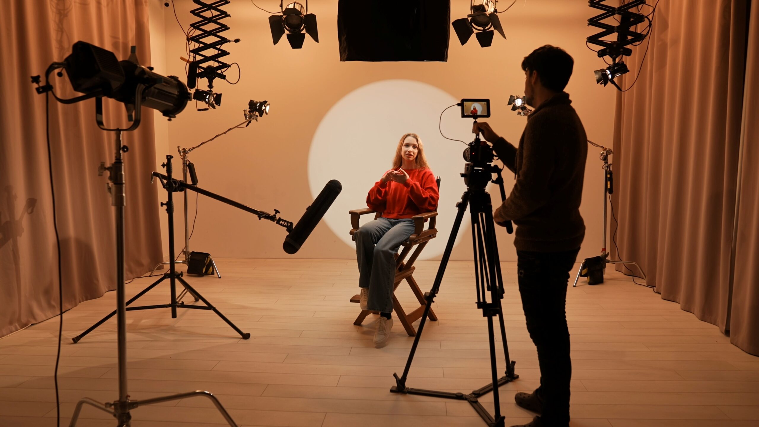 Frau sitzt im Studio auf Regiestuhl vor Kamera und Lichtsetup, während eine Person sie bei einer Filmaufnahme filmt