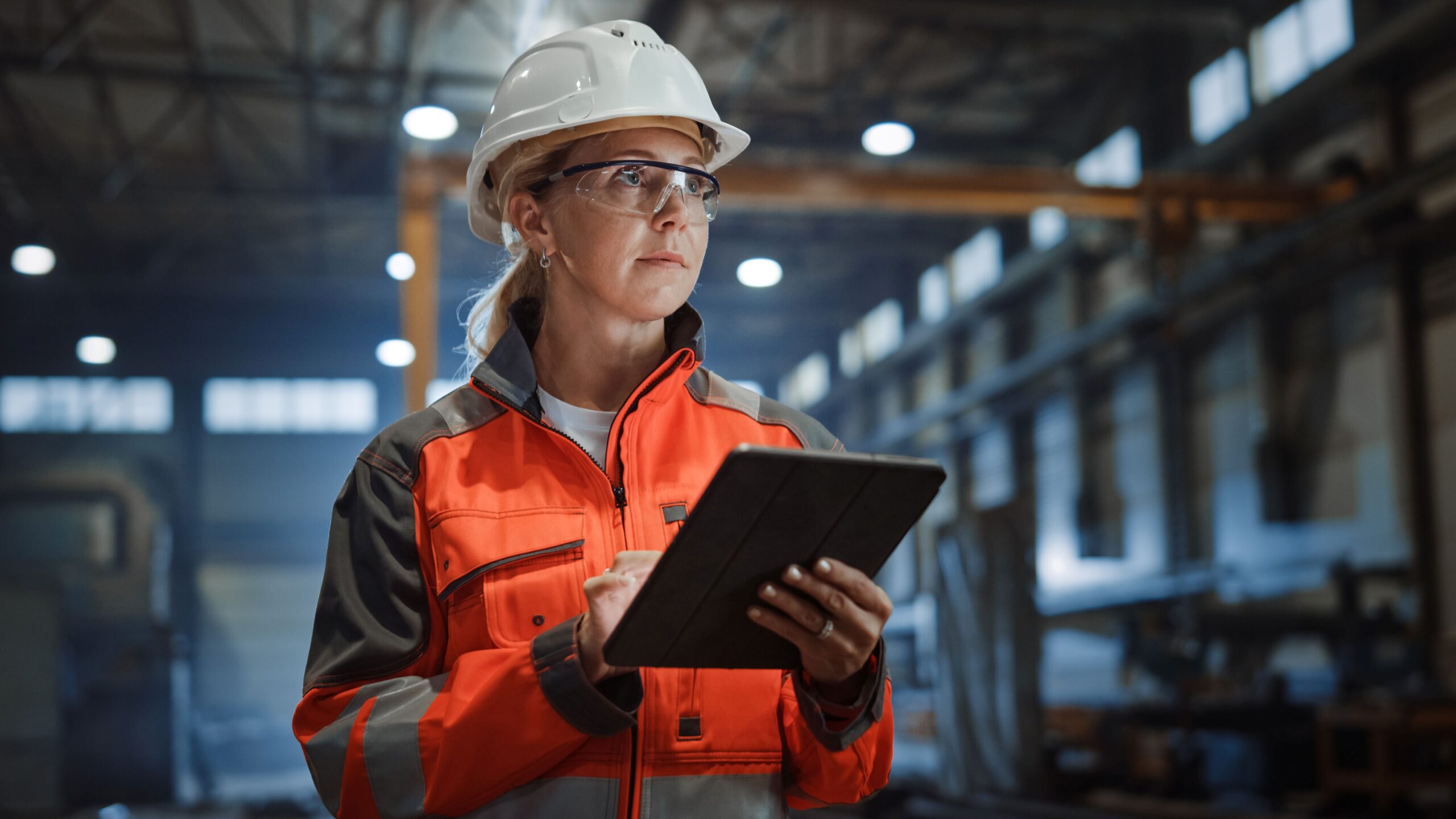 Ingenieurin überwacht Sicherheitsaspekte bei der Kabelkonfektion in einer Industriehalle mit Tablet und Schutzausrüstung