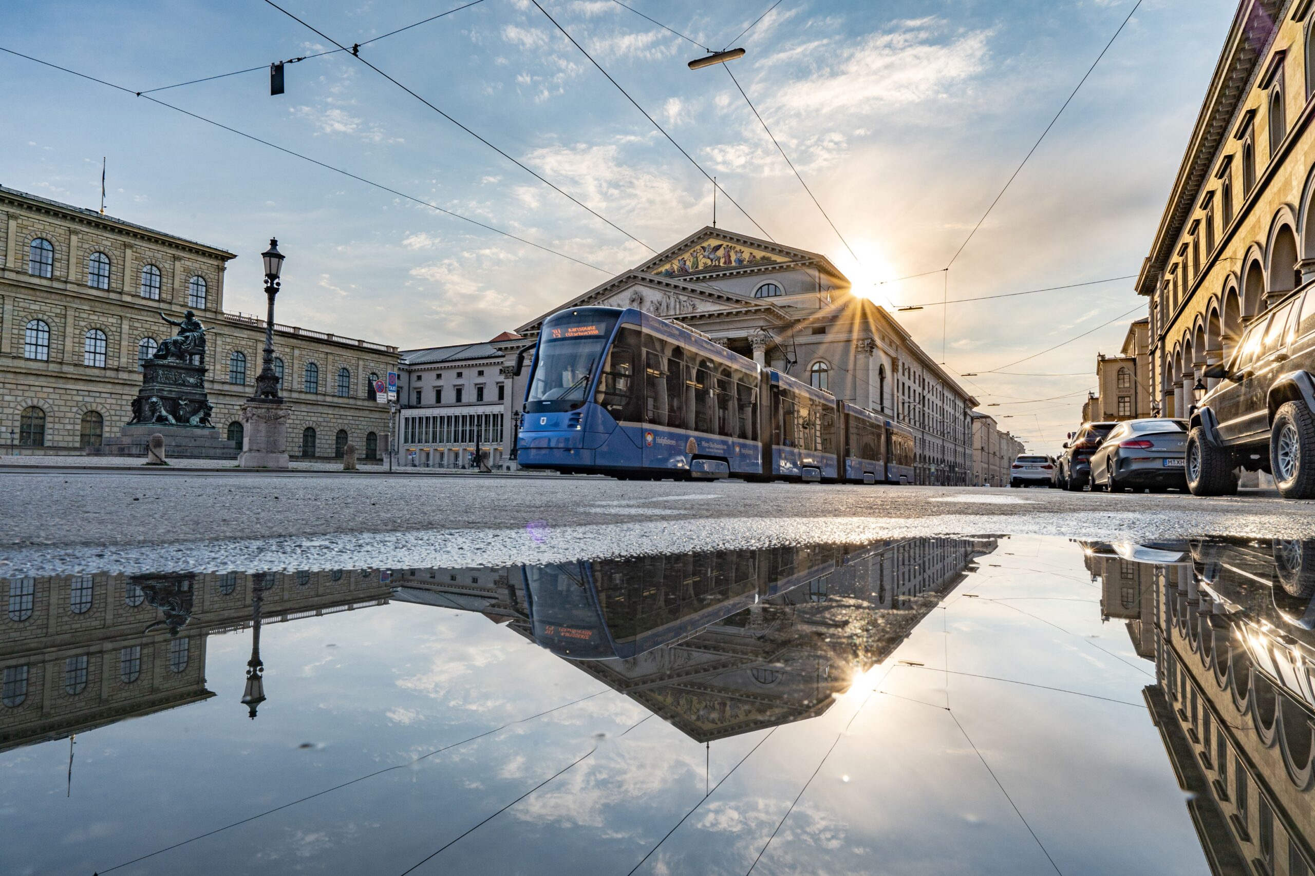 Münchner Tram vor dem Nationaltheater im Morgenlicht mit Spiegelung in Pfütze, Sinnbild für bewegende Klassenfahrt-Momente