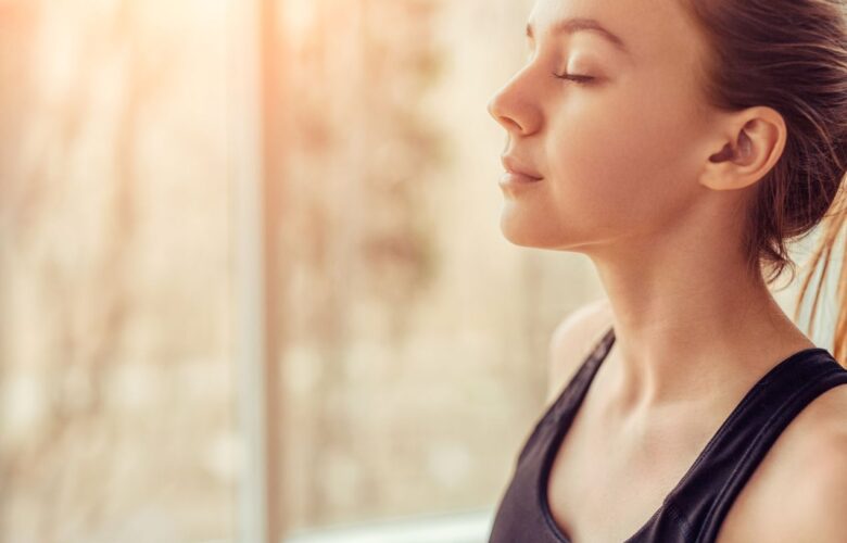 Junge Frau mit geschlossenen Augen steht im Sonnenlicht und atmet tief ein, Symbol fuer Achtsamkeit und Was ist Breathwork