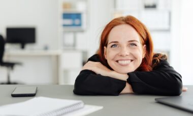 Zufriedene Frau mit rotem Haar sitzt lächelnd am Arbeitsplatz | Pflegedienst Frankfurt Stellenangebote